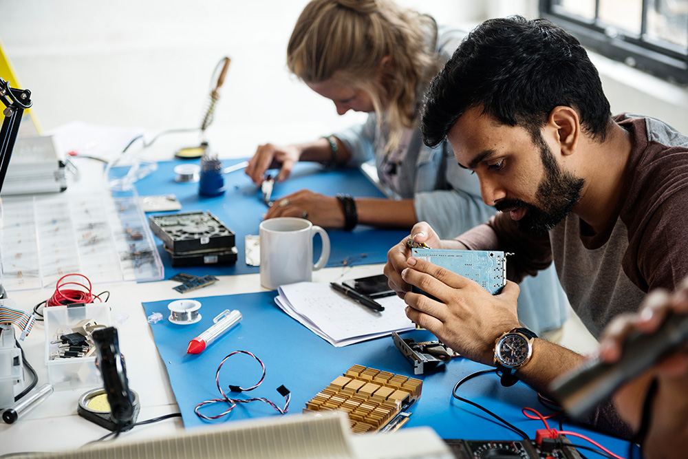 En kvinno och en man sitter vid ett bord och jobbar med elektronik.